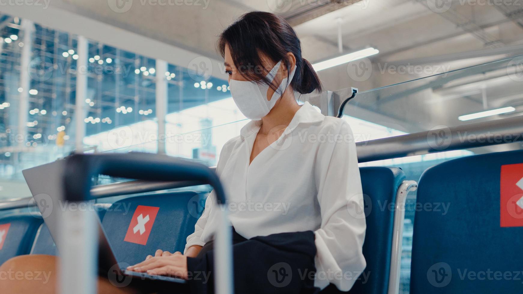 Asian business lady traveller wear face mask sitting in bench use laptop for work between wait for flight in terminal at airport. Business travel commuter in covid pandemic, Business travel concept. photo
