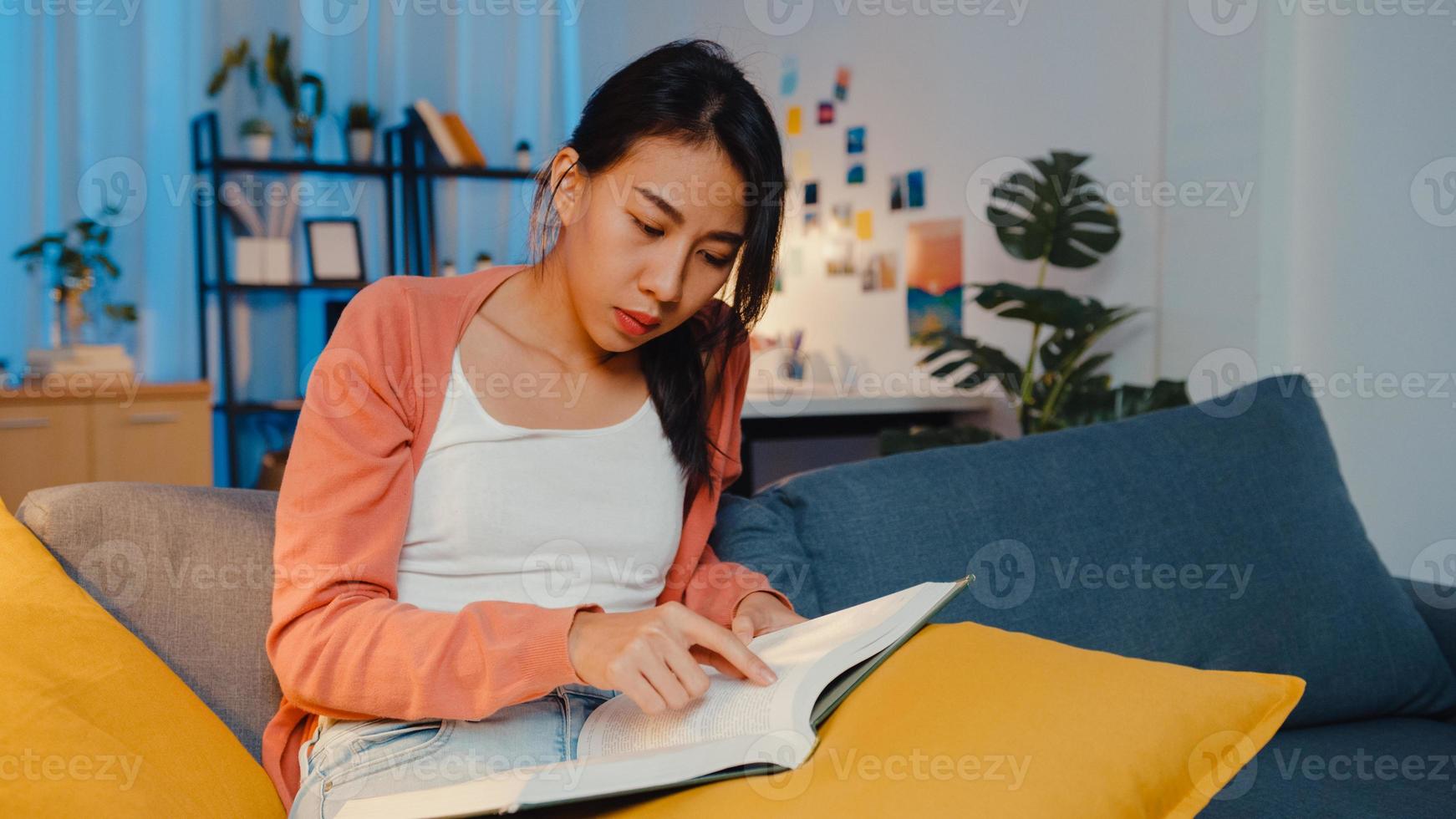 noche hermosa dama de asia lee el libro con felicidad en la tranquila sala de estar en el sofá. educación desde casa, quedarse en casa, actividad de auto cuarentena, actividad divertida para la cuarentena de covid o coronavirus. foto