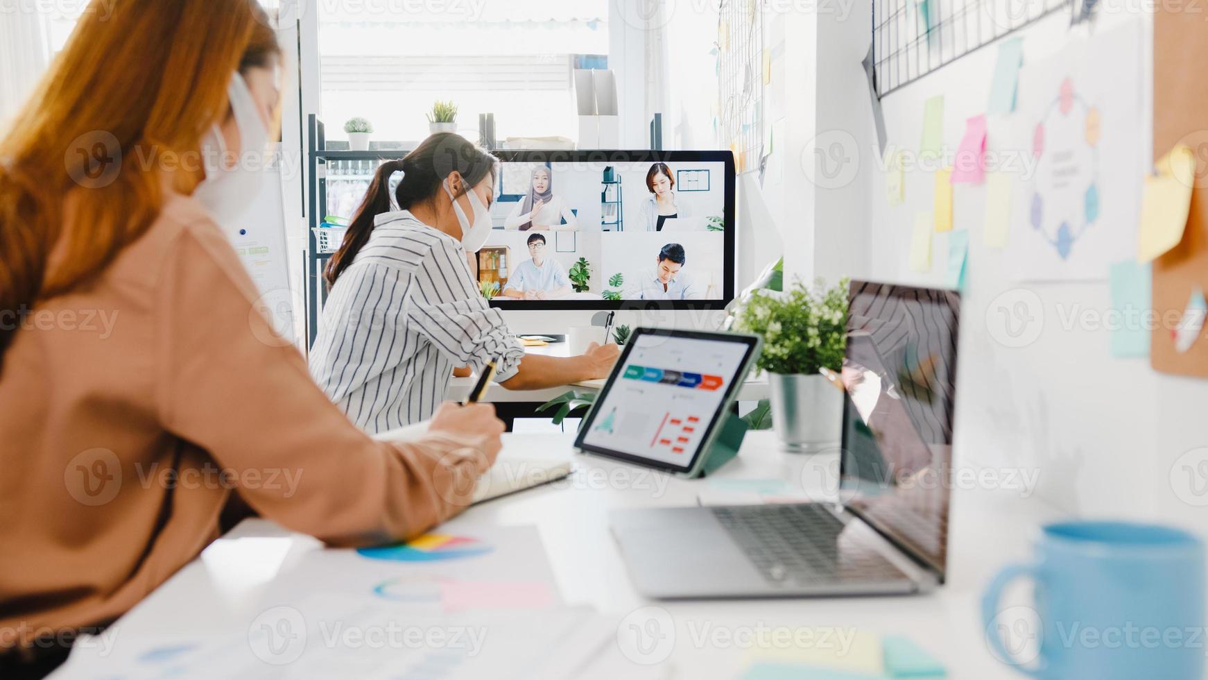 Asia businesspeople wear face mask using desktop talk to colleagues discussing brainstorm about plan in video call meeting in new normal office. Lifestyle social distancing and work after coronavirus. photo