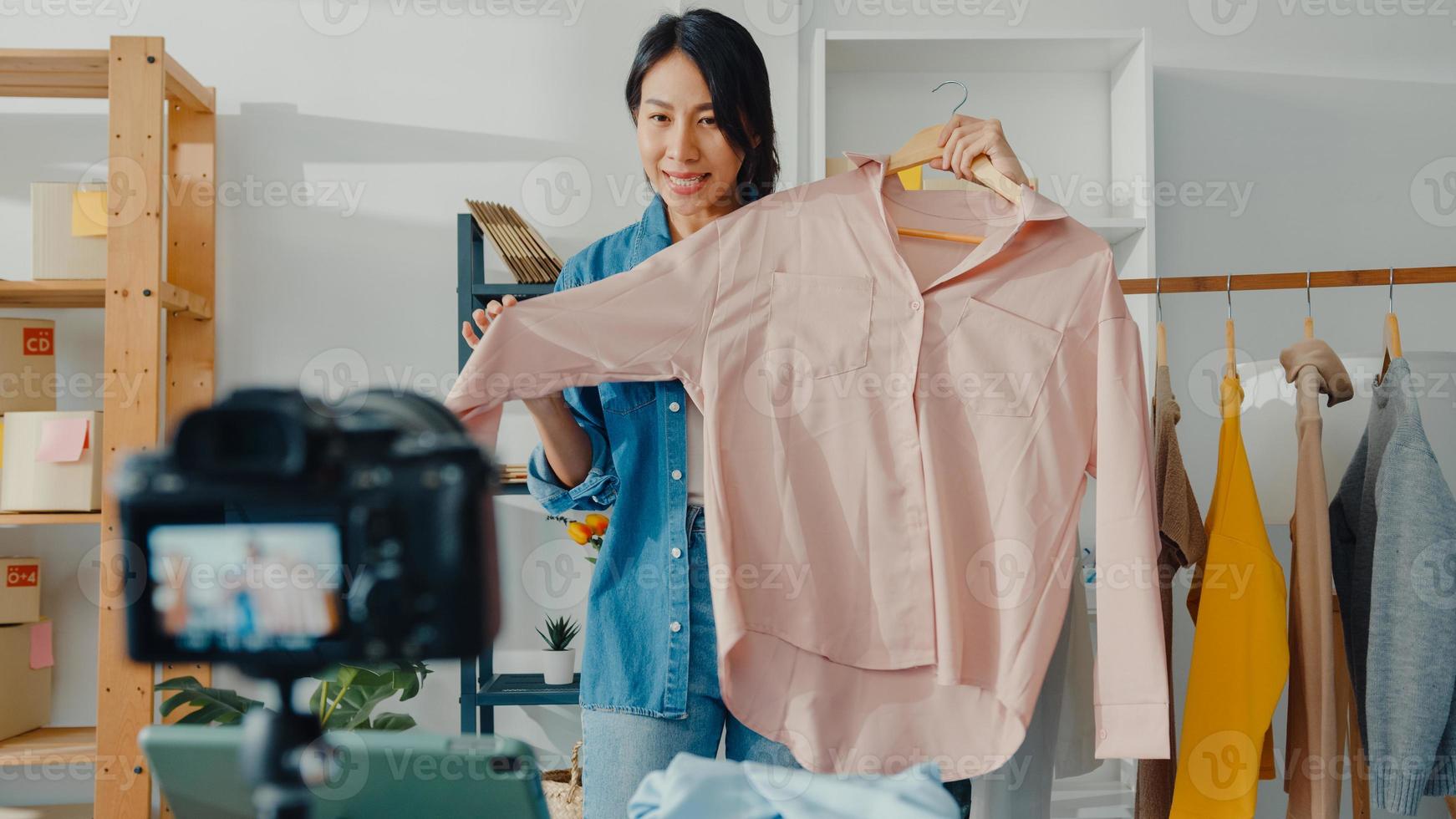 Young Asia lady fashion designer using mobile phone receiving purchase order and show clothes recording video live streaming online with camera. Small business owner, online market delivery concept. photo