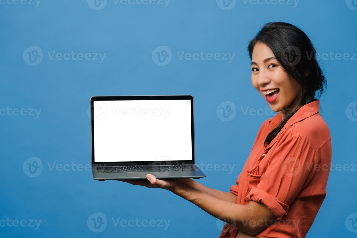 Joven asiática muestra una pantalla de computadora portátil vacía con expresión positiva, sonríe ampliamente, vestida con ropa casual sintiendo felicidad aislada sobre fondo azul. computadora con pantalla blanca en mano femenina. foto