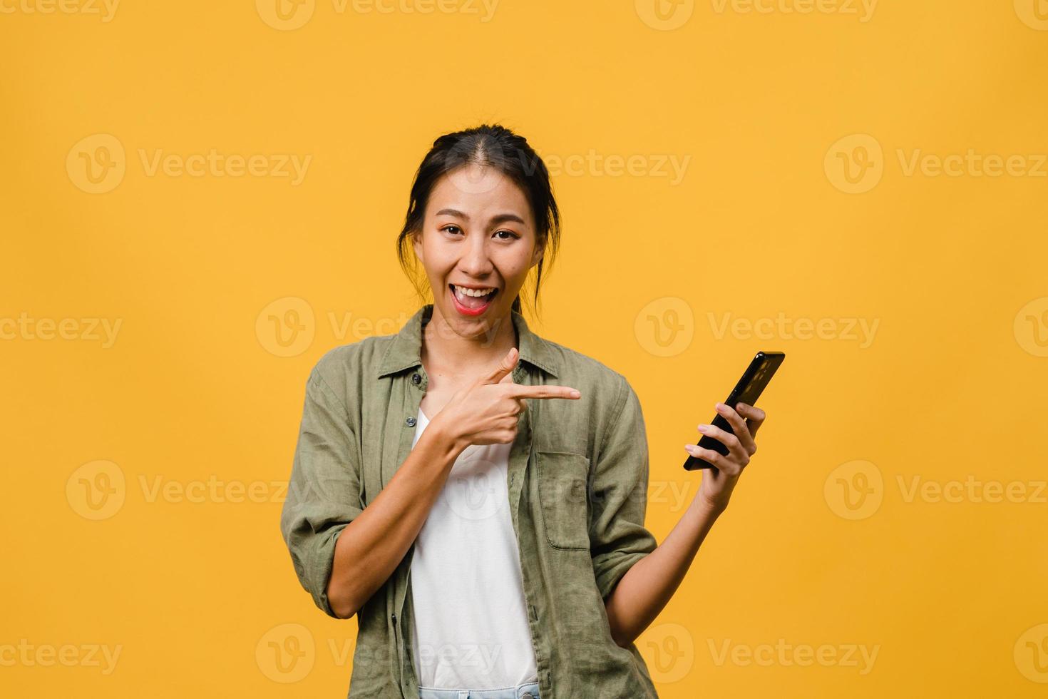 joven asiática que usa el teléfono móvil con expresión alegre, muestra algo sorprendente en el espacio en blanco en un paño casual y mirando a la cámara aislada sobre fondo amarillo. concepto de expresión facial. foto