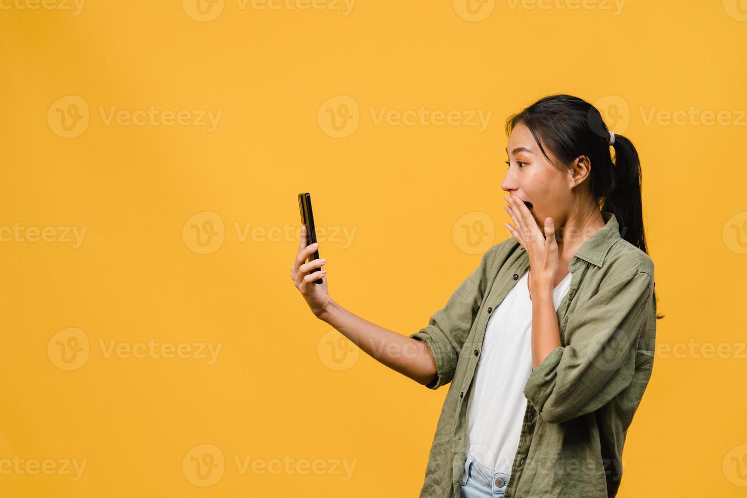 Jovencita asiática que usa el teléfono con expresión positiva, sonríe ampliamente, vestida con ropa casual sintiendo felicidad y parada aislada sobre fondo amarillo. feliz adorable mujer alegre se regocija con el éxito. foto