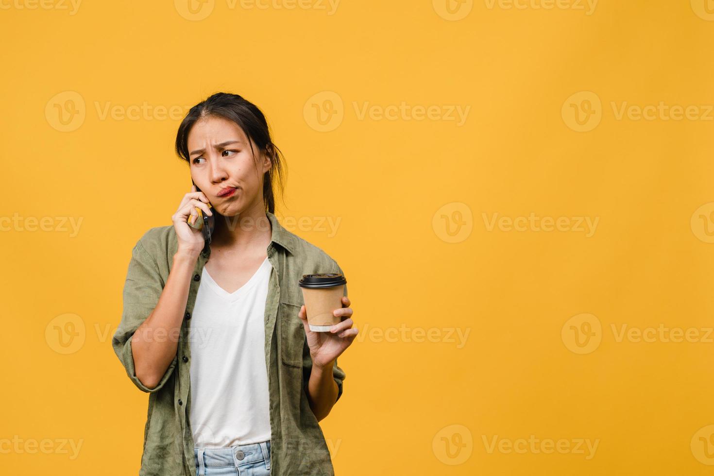 Young Asia lady talk by phone and hold coffee cup with negative expression, excited screaming, cry emotional angry in casual cloth and stand isolated on yellow background. Facial expression concept. photo