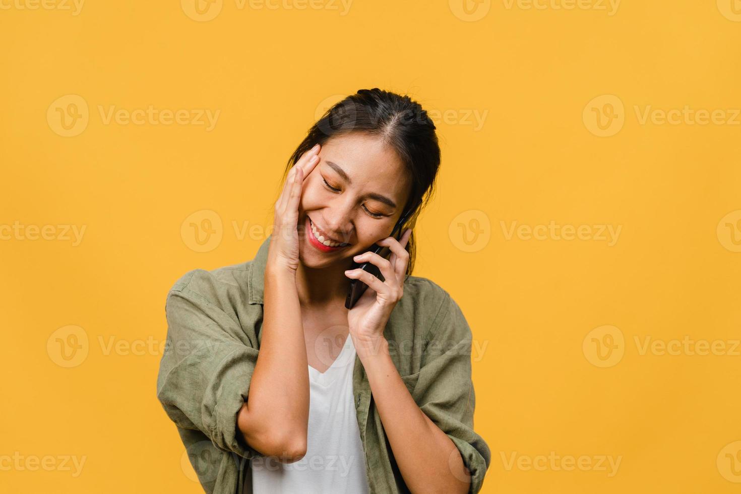 Jovencita asiática habla por teléfono con expresión positiva, sonríe ampliamente, vestida con ropa informal sintiendo felicidad y parada aislada sobre fondo amarillo. feliz adorable mujer alegre se regocija con el éxito. foto