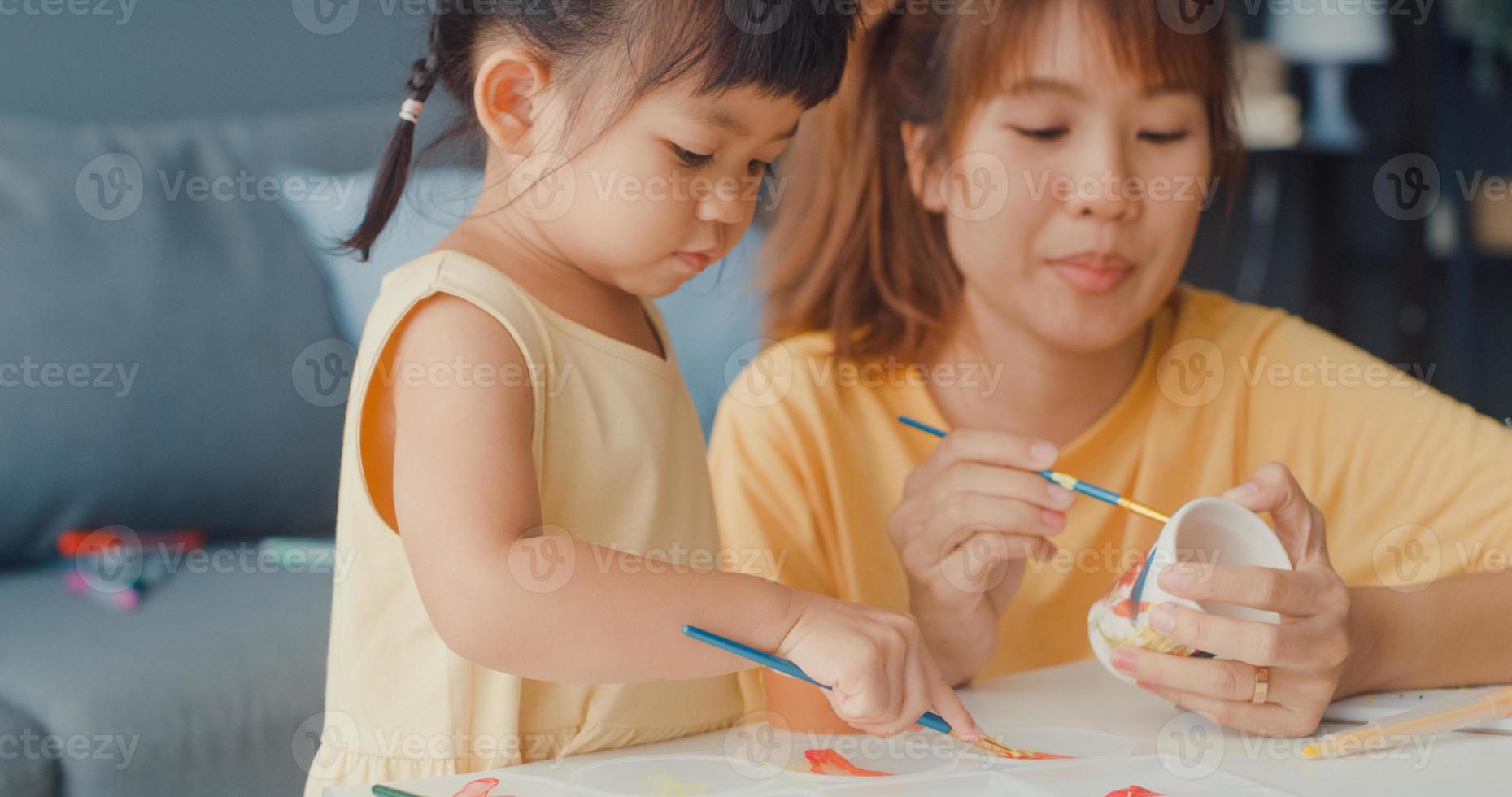 Happy cheerful Asia family mom teach toddler girl paint ceramic pot having fun relax on table in living room at house. Spending time together, Social distance, Quarantine for coronavirus prevention. photo