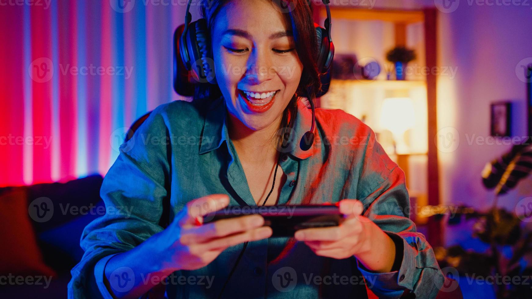 Happy asia girl gamer wear headphone competition play video game online with smartphone colorful neon lights in living room at home. Esport streaming game online, Home quarantine activity concept. photo