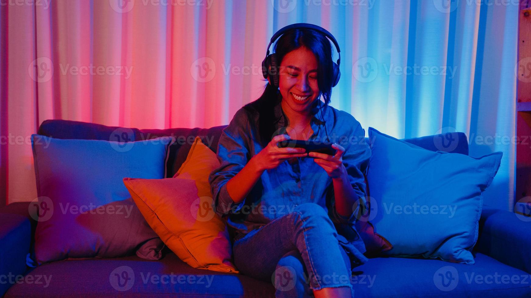Happy asia girl gamer wear headphone competition video game online with smartphone excited talk with friend sit on couch in colorful neon lights living room at home, Home quarantine activity concept. photo