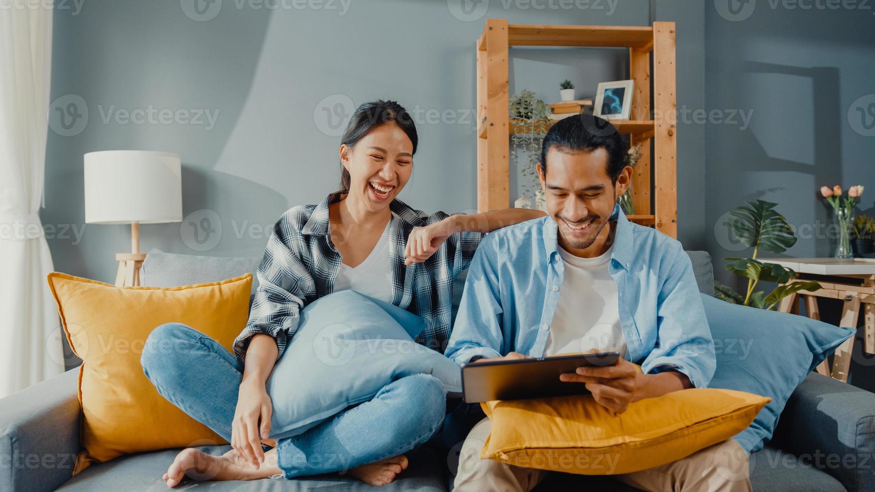 Feliz pareja joven y atractiva asiática, hombre y mujer, sentarse en el sofá, usar la tableta, comprar muebles en línea, decorar el hogar en la sala de estar de la nueva casa. joven casado mudanza concepto online comprador de casa. foto