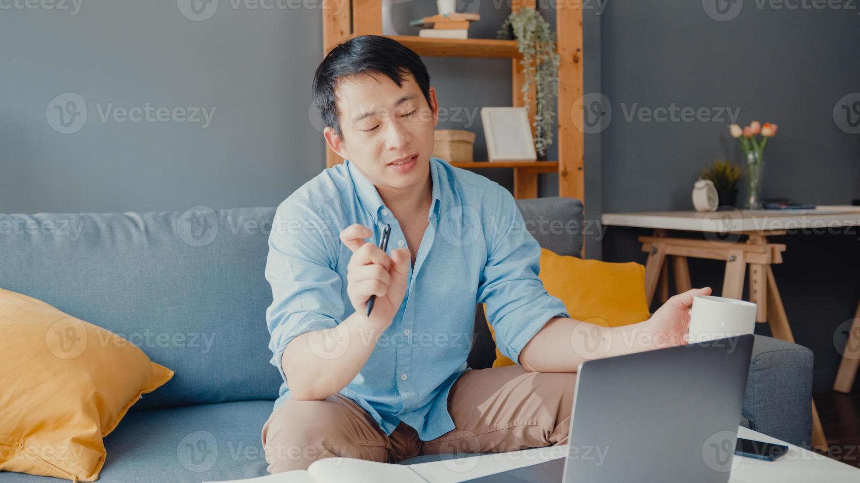 Young Asia businessman using laptop talk to colleagues about plan in video call while smart working from home at living room. Self-isolation, social distancing, quarantine for corona virus prevention. photo