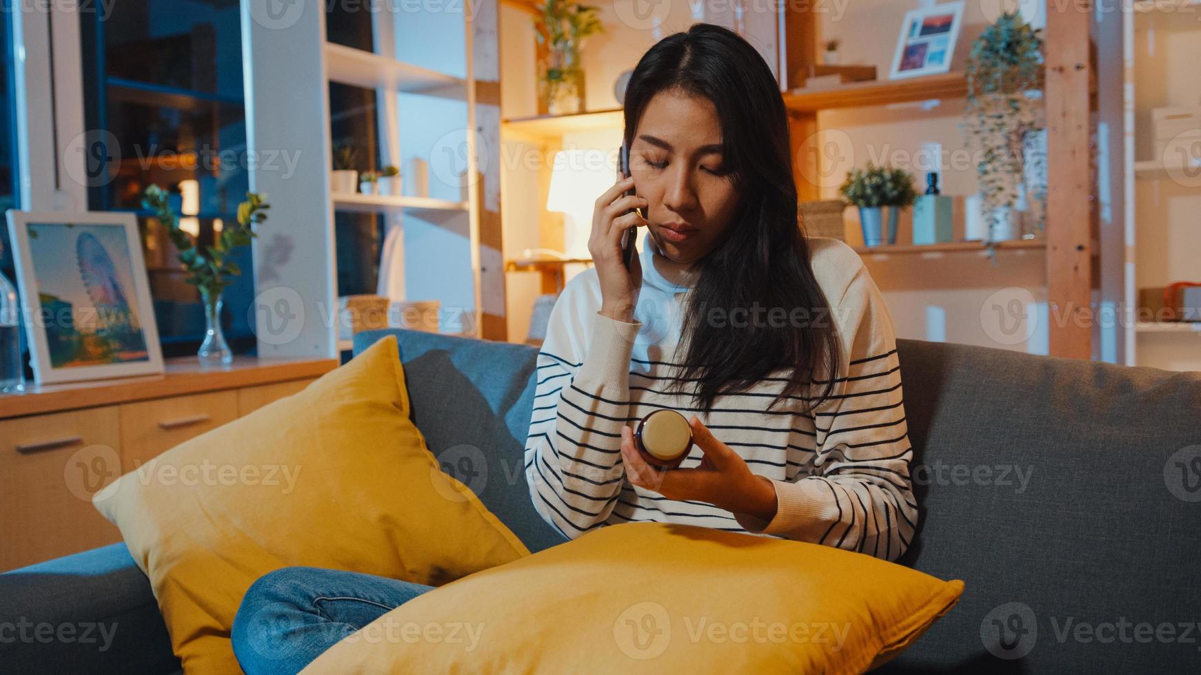 Sick young Asian woman hold medicine sit on couch use smart phone to consult with doctor at home night. Girl take medicine after doctor order, quarantine at house, Social distance coronavirus concept. photo