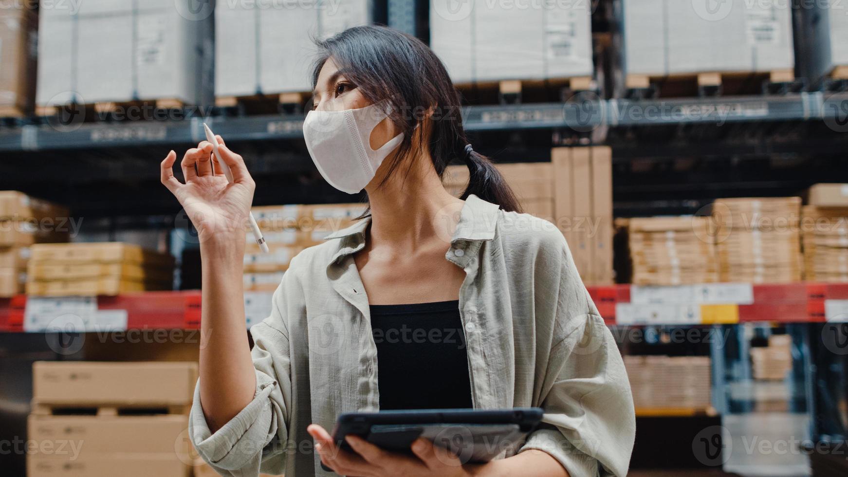 Young Asia businesswoman manager wear face mask looking for goods using digital tablet checking inventory levels stand in retail shopping center. Distribution, Logistics, Packages ready for shipment. photo