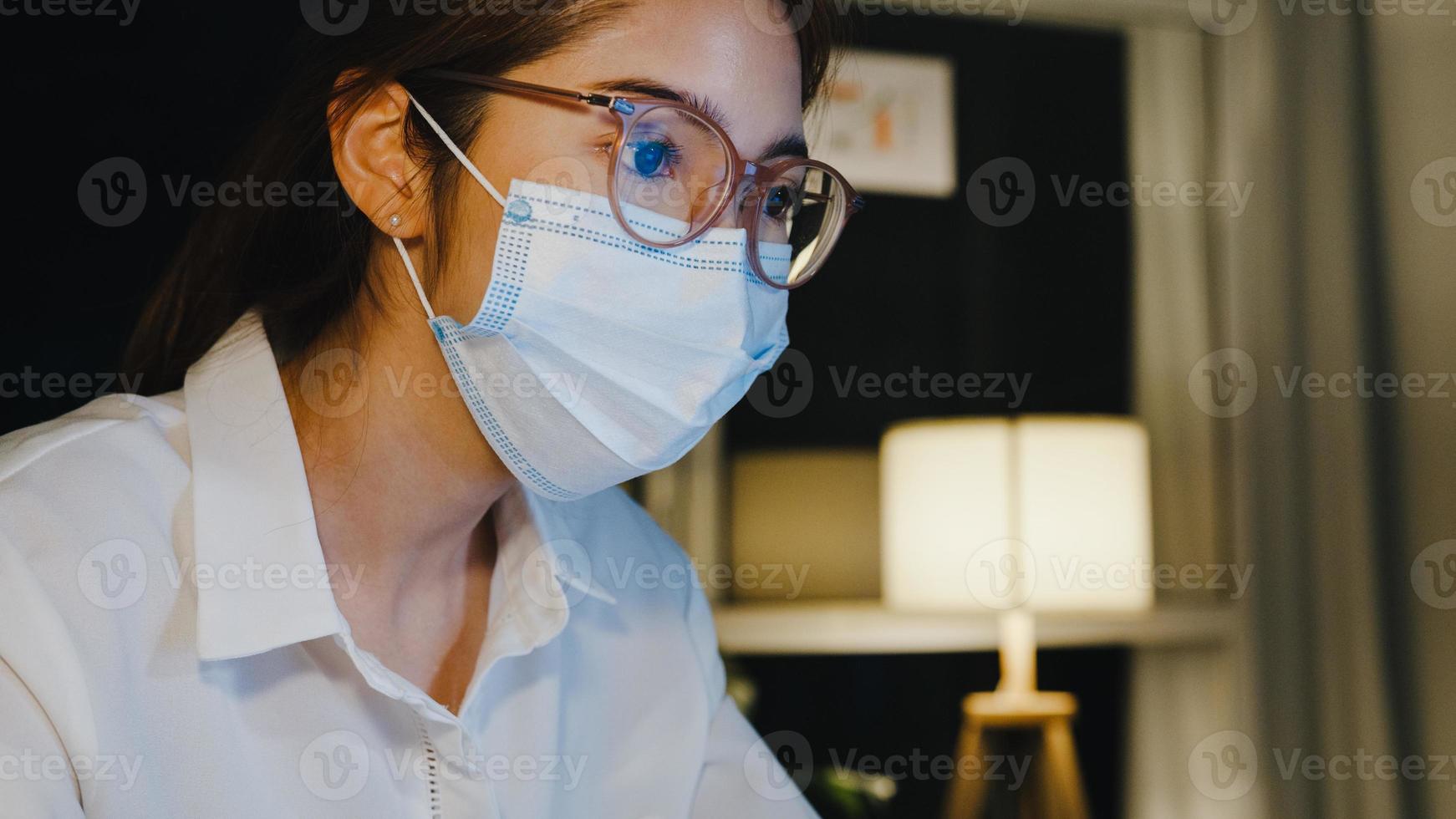 Freelance Asia lady wear medical face mask use laptop hard work in living room at house. Working from home overload at night, remotely work, social distancing, quarantine for corona virus prevention. photo