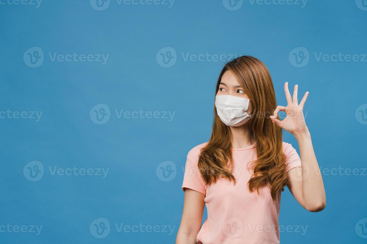 Young Asia girl wearing medical face mask gesturing ok sign with dressed in casual cloth and look at camera isolated on blue background. Self-isolation, social distancing, quarantine for corona virus. photo