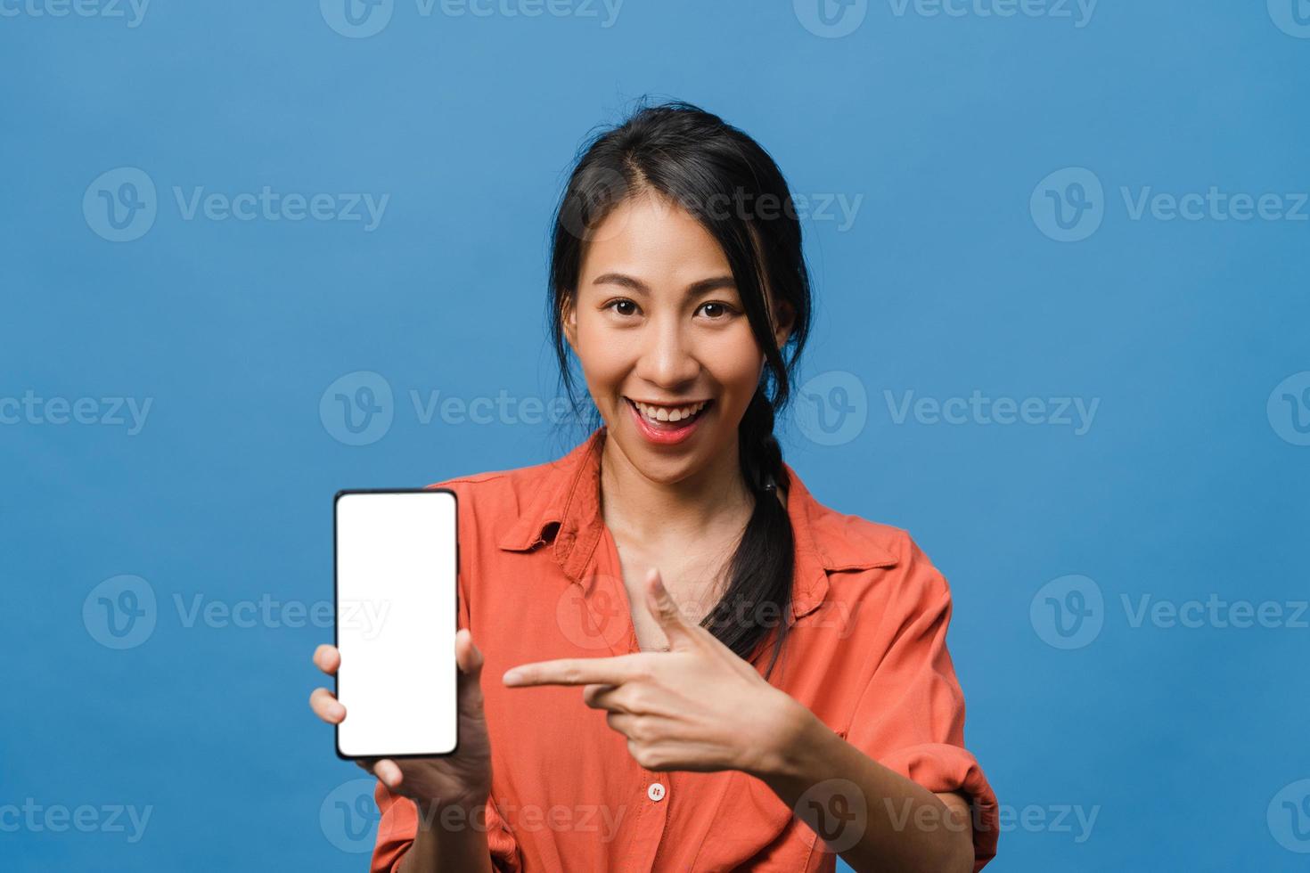 Joven asiática muestra la pantalla vacía del teléfono inteligente con expresión positiva, sonríe ampliamente, vestida con ropa casual sintiendo felicidad sobre fondo azul. teléfono móvil con pantalla en blanco en mano femenina. foto