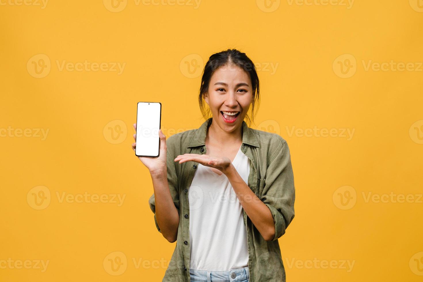 Joven asiática muestra la pantalla vacía del teléfono inteligente con expresión positiva, sonríe ampliamente, vestida con ropa casual sintiendo felicidad sobre fondo amarillo. teléfono móvil con pantalla en blanco en mano femenina. foto