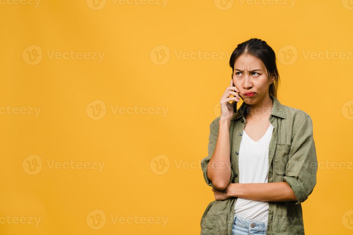 jovencita asiática habla por teléfono con expresión negativa, gritos emocionados, gritos emocionales enojados en ropa casual y se para aislado sobre fondo amarillo con espacio de copia en blanco. concepto de expresión facial. foto
