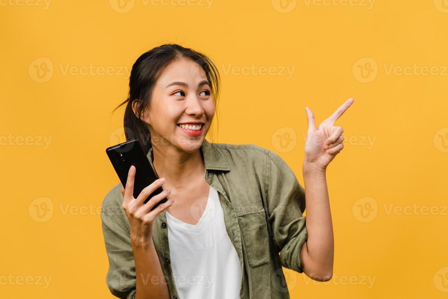 Portrait of young Asian lady using mobile phone with cheerful expression, show something amazing at blank space in casual clothing and stand isolated over yellow background. Facial expression concept. photo