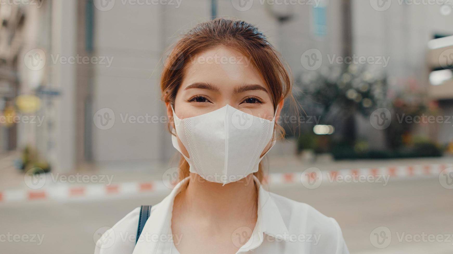 Successful young Asia businesswoman in fashion office clothes wear medical face mask smiling and looking at camera while happy standing alone outdoors in urban modern city. Business on the go concept. photo
