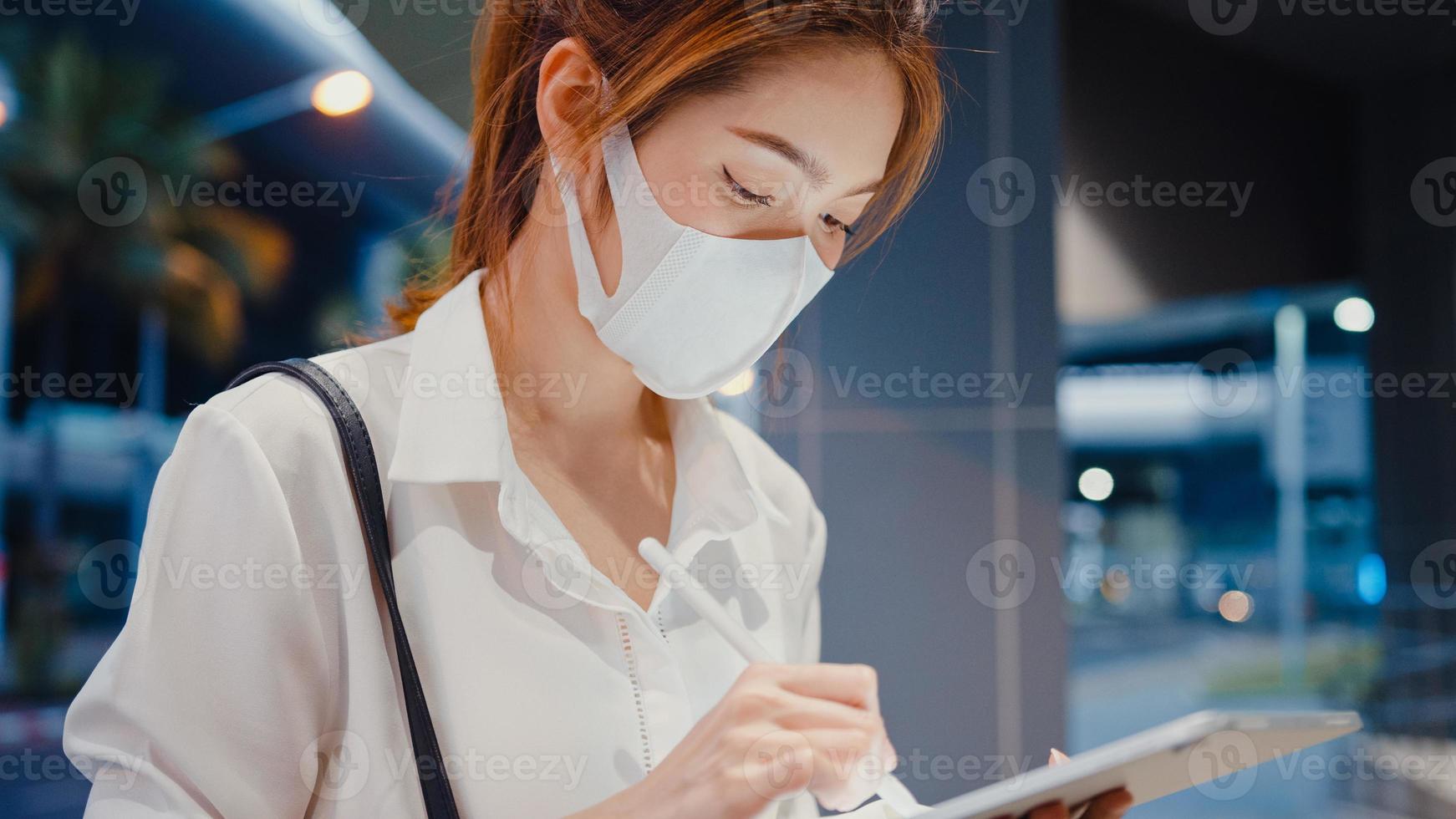 Joven empresaria asiática en ropa de oficina de moda usa mascarilla médica con bolígrafo inteligente para escribir en tableta digital mientras se sienta sola al aire libre en la ciudad moderna urbana por la noche. concepto de negocio en movimiento. foto