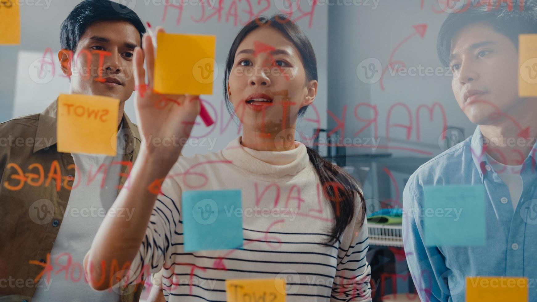 Asia businesspeople stand behind transparent glass wall listen manager pointing progress work and brainstorm meeting and worker post sticky note on wall at office. Business inspiration, Share ideas. photo