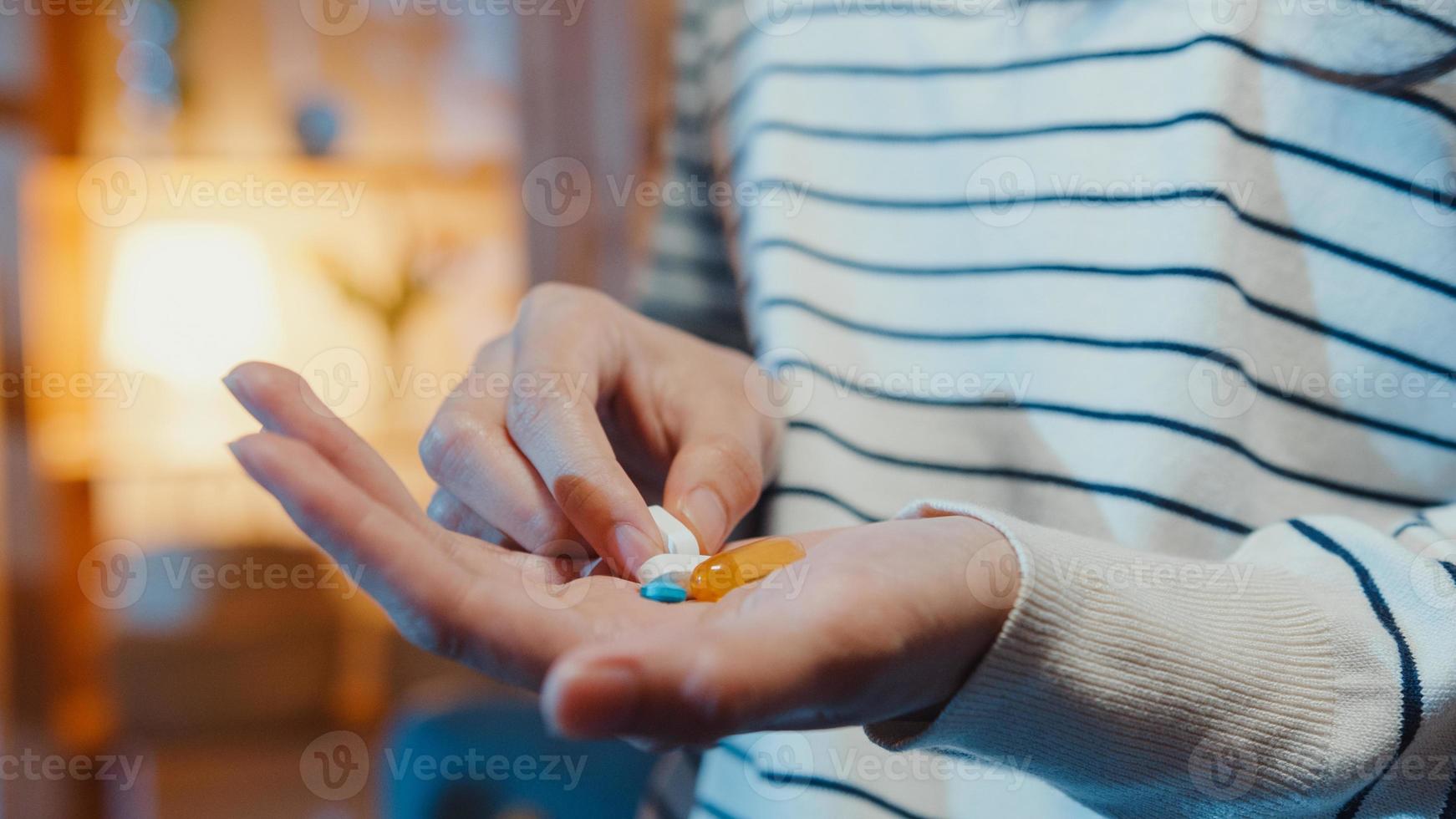 Sick asian young lady holding pill take medicine sit on couch in living room at home night. Girl taking drug after doctor order, quarantine at house, Coronavirus social distancing healthcare concept. photo