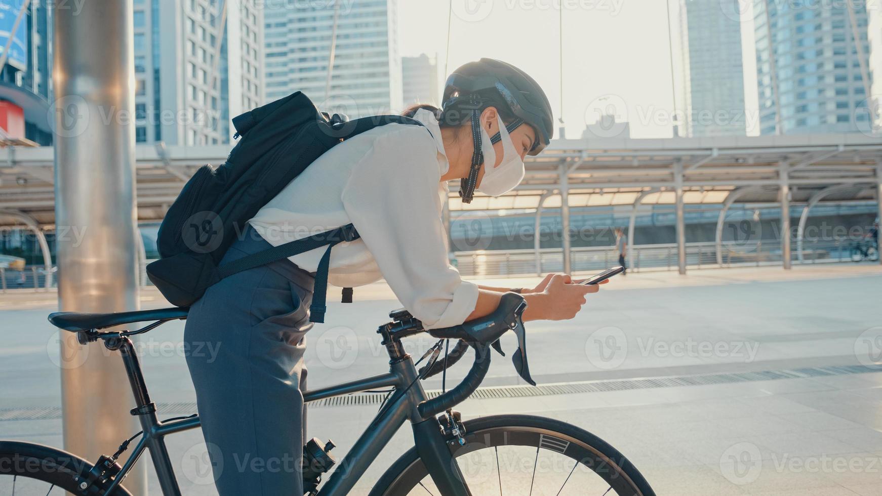 Asian businesswoman carry backpack wear anti virus protection mask take bicycle walk and check phone in city street go to work at office. commute to work, Business commuter for covid-19 concept. photo