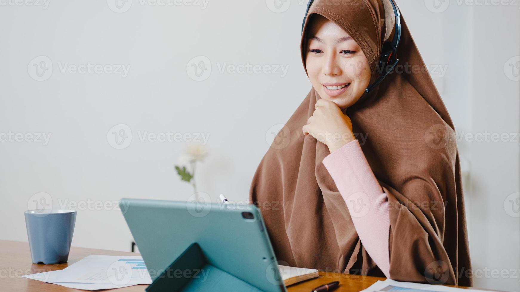 Asia dama musulmana usa auriculares con tableta digital, hable con sus colegas sobre el informe de venta en una videollamada de conferencia mientras trabaja desde casa en la cocina. distanciamiento social, cuarentena por coronavirus. foto