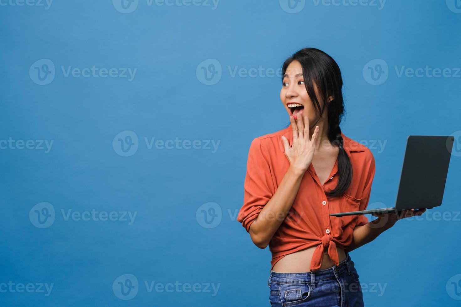 Jovencita asiática usando laptop con expresión positiva, sonríe ampliamente, vestida con ropa casual sintiendo felicidad y parada aislada sobre fondo azul. feliz adorable mujer alegre se regocija con el éxito. foto