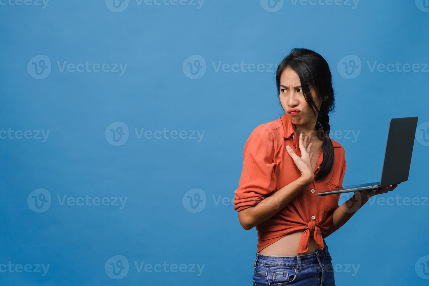 Jovencita asiática usando laptop con expresión negativa, gritando emocionado, llorando emocionalmente enojado en ropa casual y parado aislado sobre fondo azul con espacio de copia en blanco. concepto de expresión facial. foto