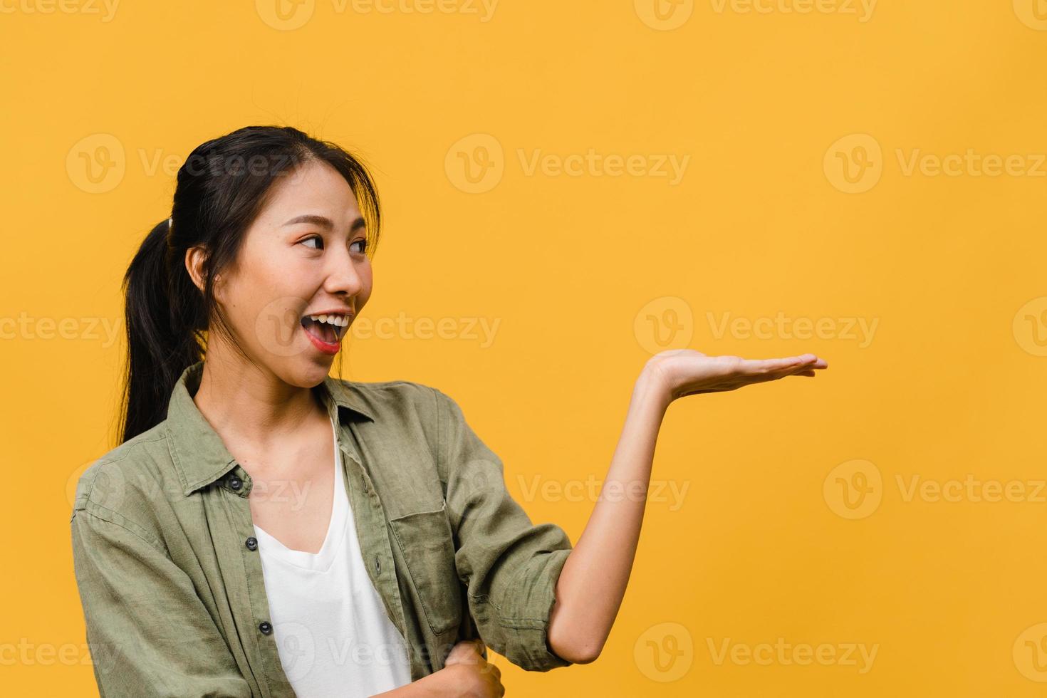 Portrait of young Asian lady smiling with cheerful expression, shows something amazing at blank space in casual clothing and standing isolated over yellow background. Facial expression concept. photo