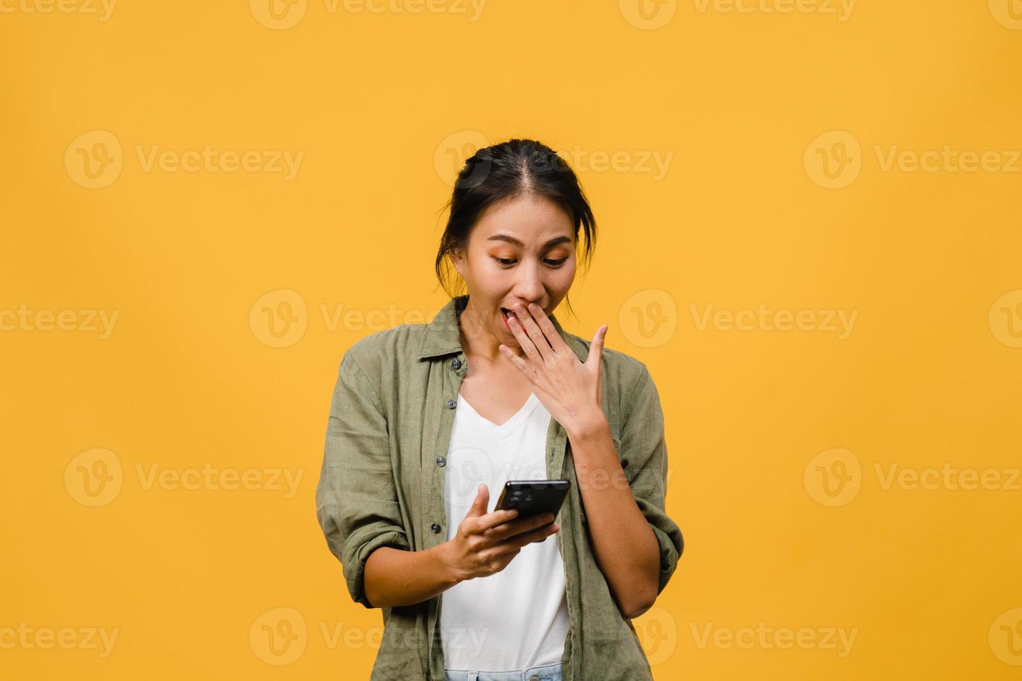 Young Asia lady using phone with positive expression, smiles broadly, dressed in casual clothing feeling happiness and stand isolated on yellow background. Happy adorable glad woman rejoices success. photo