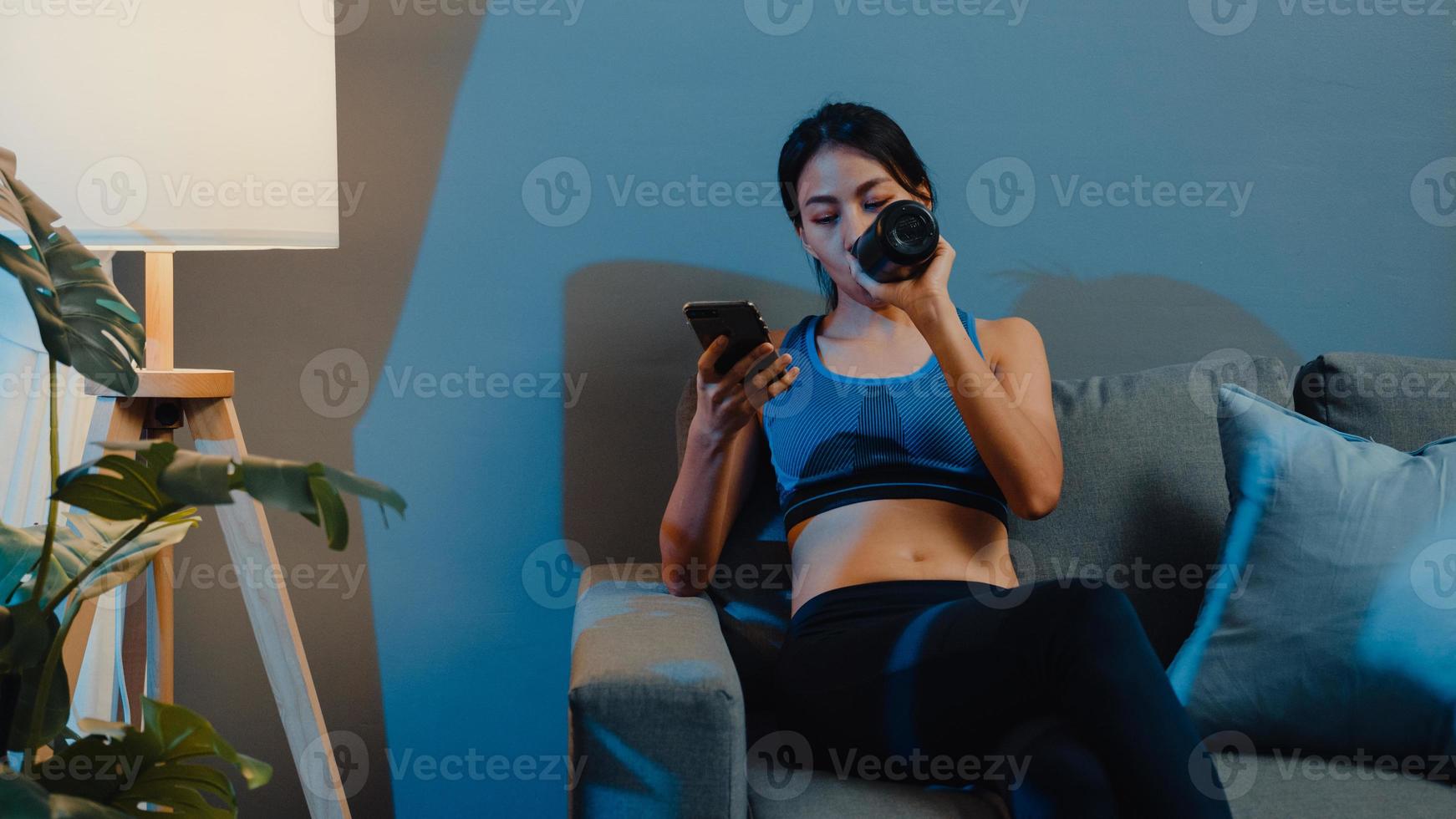 Young Asia lady using mobile phone and drinking pure water on couch in living room at home night. Sport and recreation activity, social distancing, quarantine for corona virus prevention concept. photo