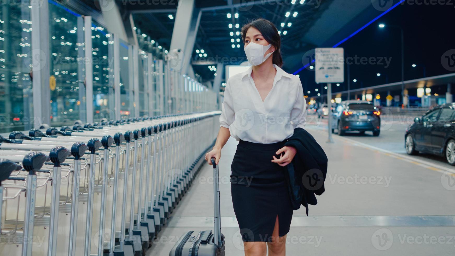 Asian business girl arrive destination wear face mask with drag luggage walk outside wait car terminal at domestic airport. Business commuter covid pandemic, Business travel social distancing concept. photo