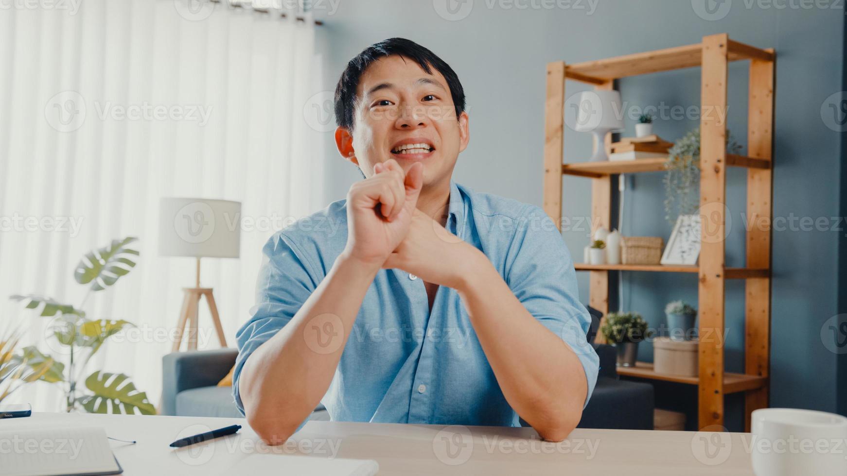Young Asia businessman using computer laptop talk to colleagues about plan in video call meeting while working from home at living room. Self-isolation, social distancing, quarantine for corona virus. photo