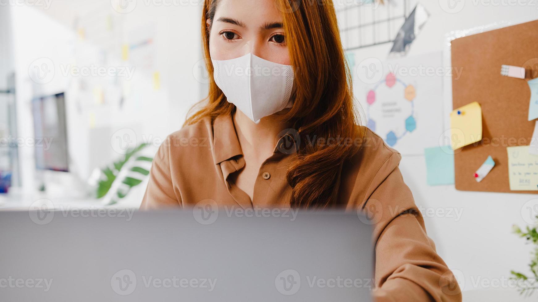 Asia businesswoman wear face mask for social distancing in new normal situation for virus prevention while using laptop presentation to colleagues about plan in video call while work in office. photo