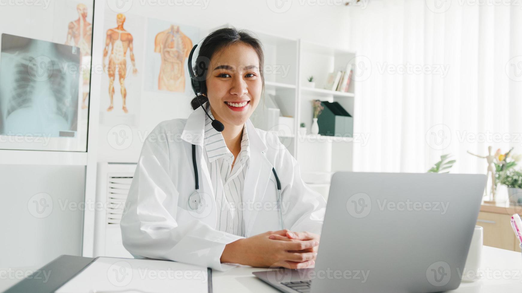 Young Asia lady doctor in white medical uniform with stethoscope using computer laptop talking video conference call with patient at desk in health clinic or hospital. Consulting and therapy concept. photo