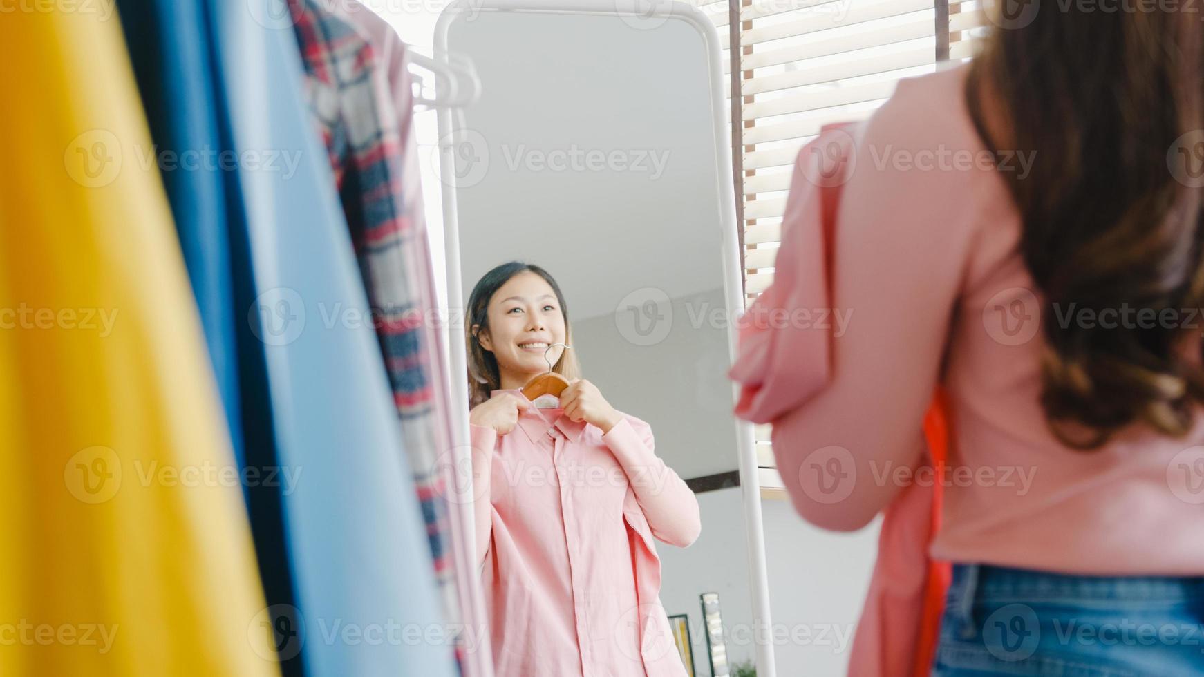 Bella dama atractiva de Asia eligiendo ropa en perchero vestirse mirando a sí misma en el espejo en el dormitorio en casa. chica piensa qué ponerse camisa casual. las mujeres de estilo de vida se relajan en el concepto de hogar. foto