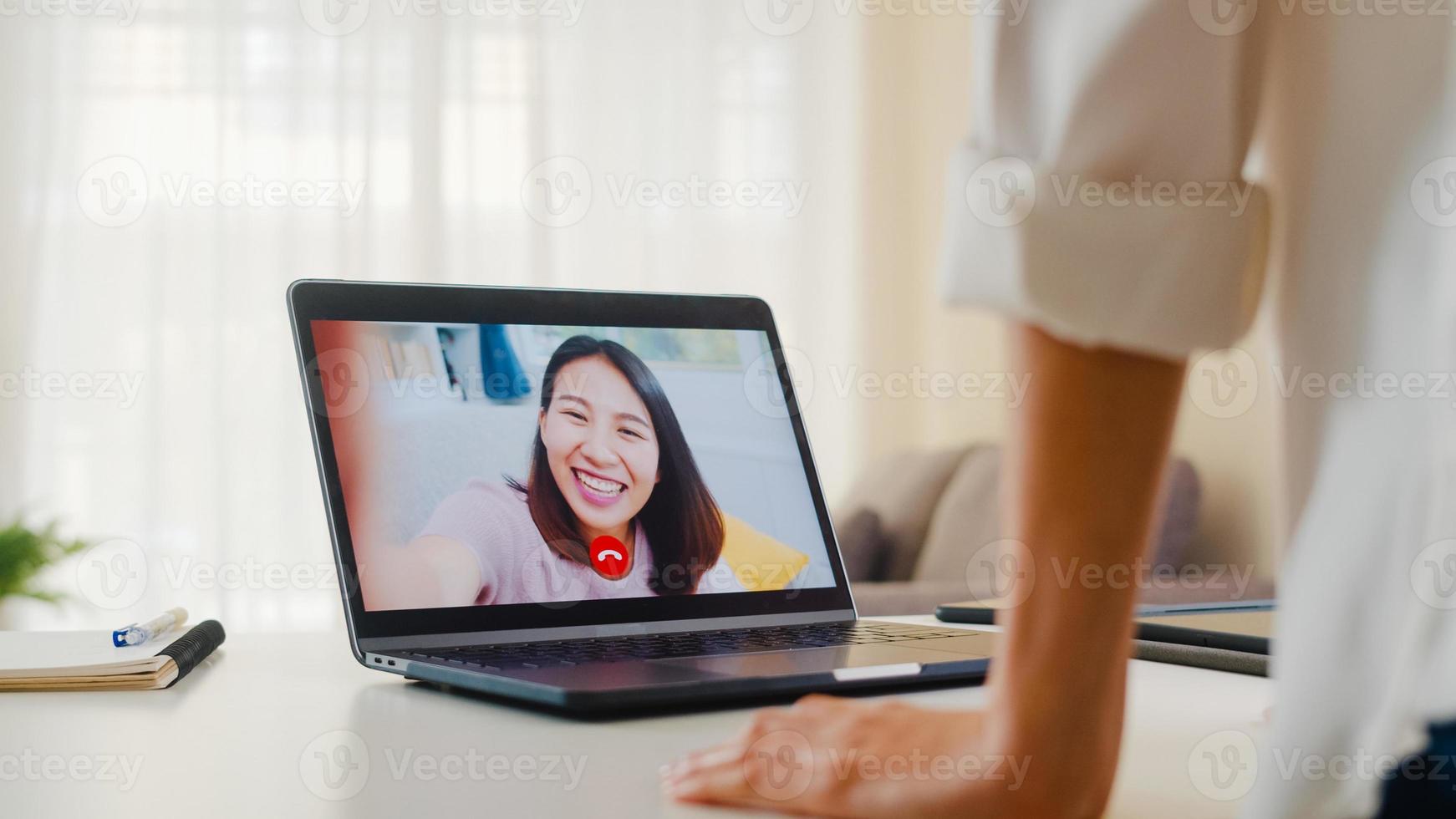 Young Asian business female using laptop video call talking with friends while working from home at living room. Self-isolation, social distancing, quarantine for coronavirus in next normal concept. photo