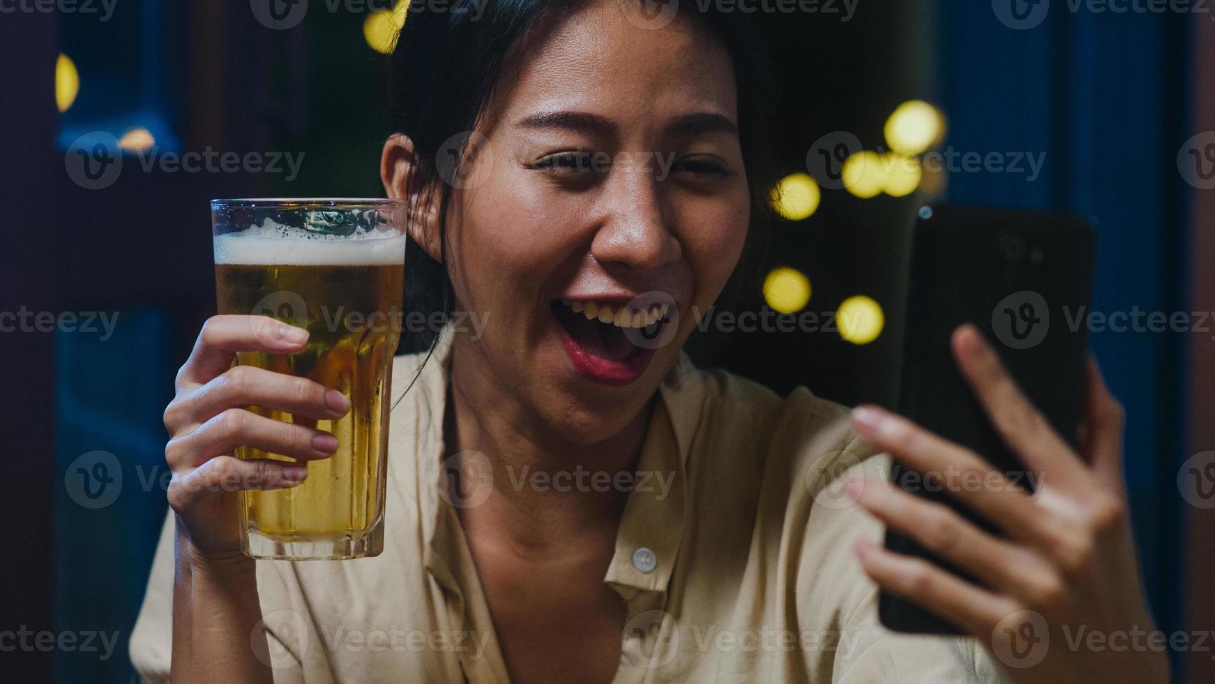 joven asiática bebiendo cerveza divirtiéndose feliz momento fiesta nocturna evento de año nuevo celebración en línea a través de videollamada por teléfono en casa por la noche. distanciamiento social, cuarentena para la prevención del coronavirus. foto