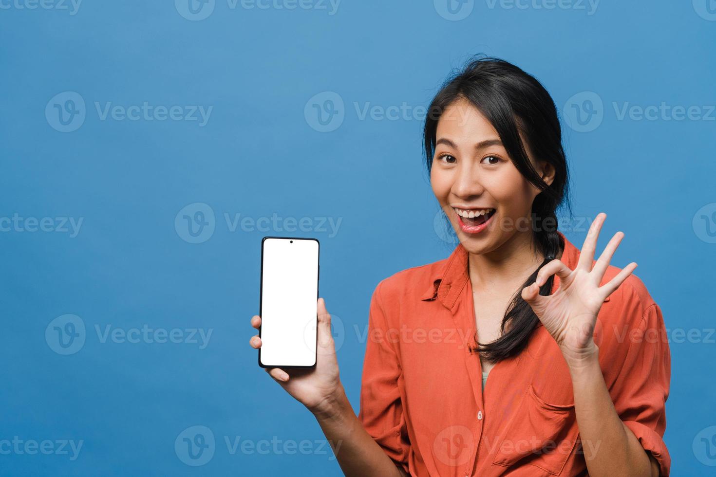 Young Asia lady show empty smartphone screen with positive expression, smiles broadly, dressed in casual clothing feeling happiness on blue background. Mobile phone with white screen in female hand. photo