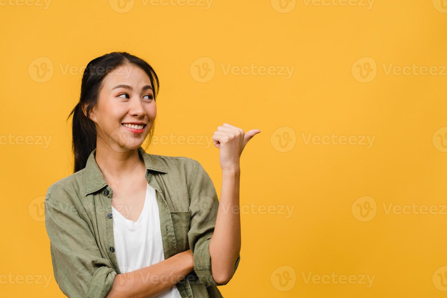 Portrait of young Asian lady smiling with cheerful expression, shows something amazing at blank space in casual clothing and standing isolated over yellow background. Facial expression concept. photo