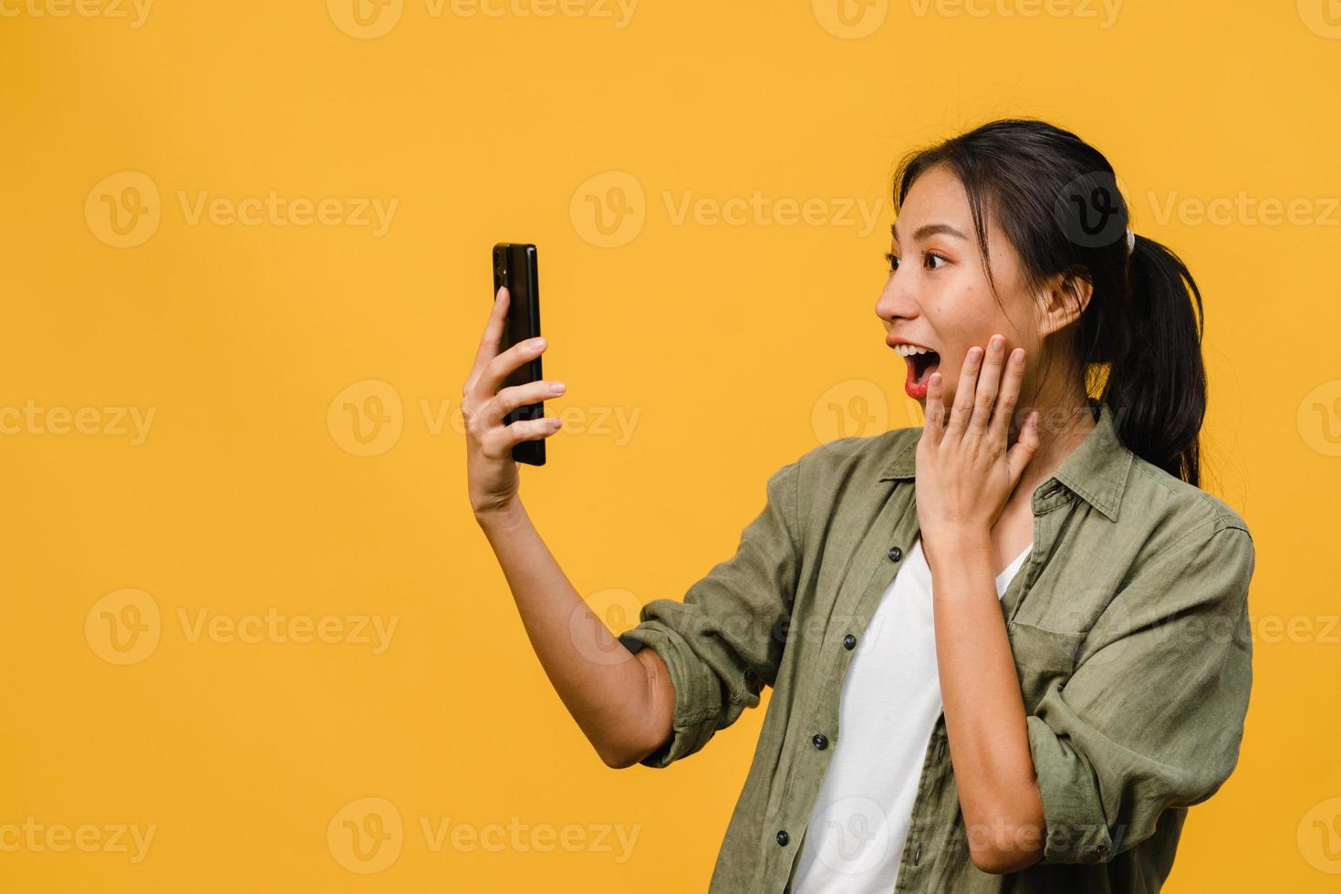 Surprised young Asia lady using mobile phone with positive expression, smiles broadly, dressed in casual clothing and stand isolated on yellow background. Happy adorable glad woman rejoices success. photo