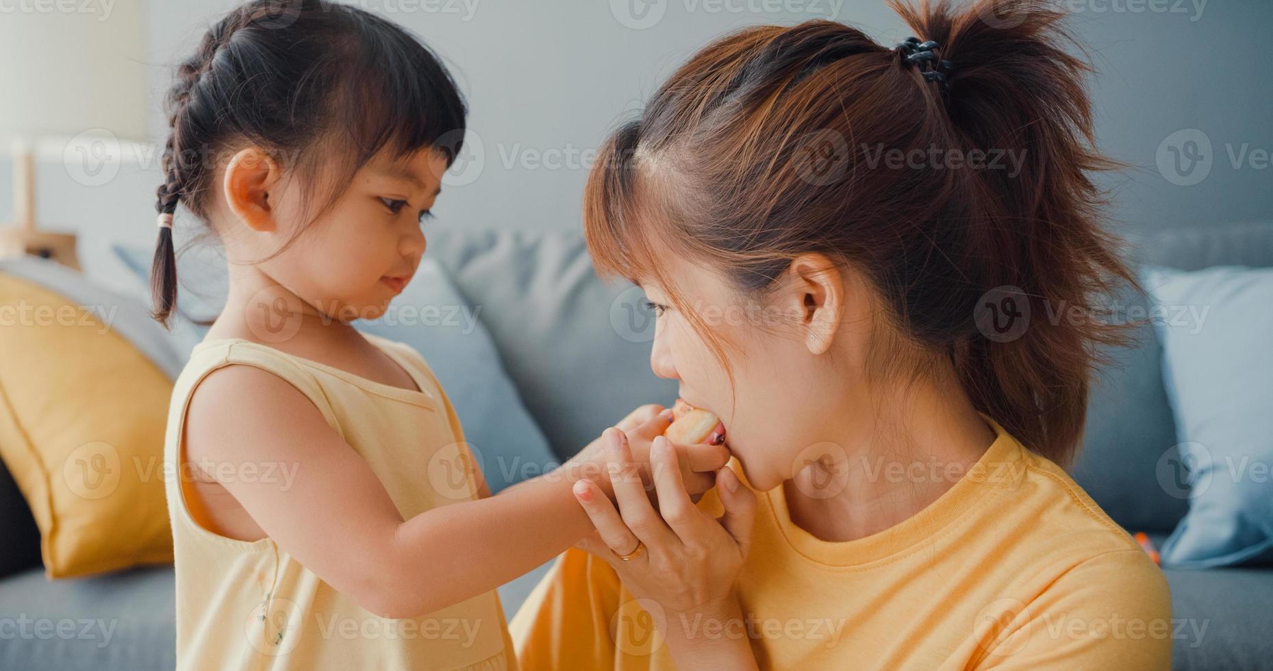 Happy cheerful Asia family mom and toddler girl eating donuts and having fun relax enjoy on couch in living room at house. Spending time together, Social distance, Quarantine for coronavirus. photo