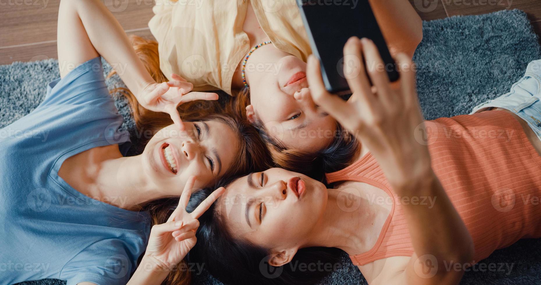 Above view close up group of Asia ladies with happiness enjoy moment hold smartphone smiling take memories picture lying on carpet in floor living room at home. Lifestyle activity quarantine concept. photo