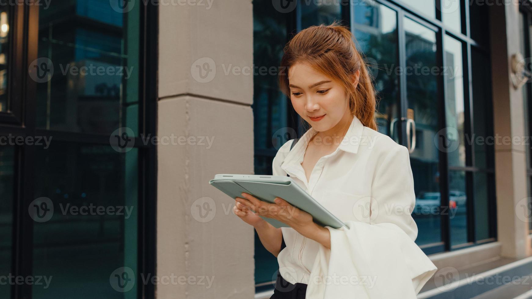 Exitosa empresaria asiática joven en ropa de oficina de moda usando tableta digital y escribiendo mensajes de texto mientras camina sola al aire libre en la ciudad moderna urbana por la mañana. concepto de negocio en movimiento. foto