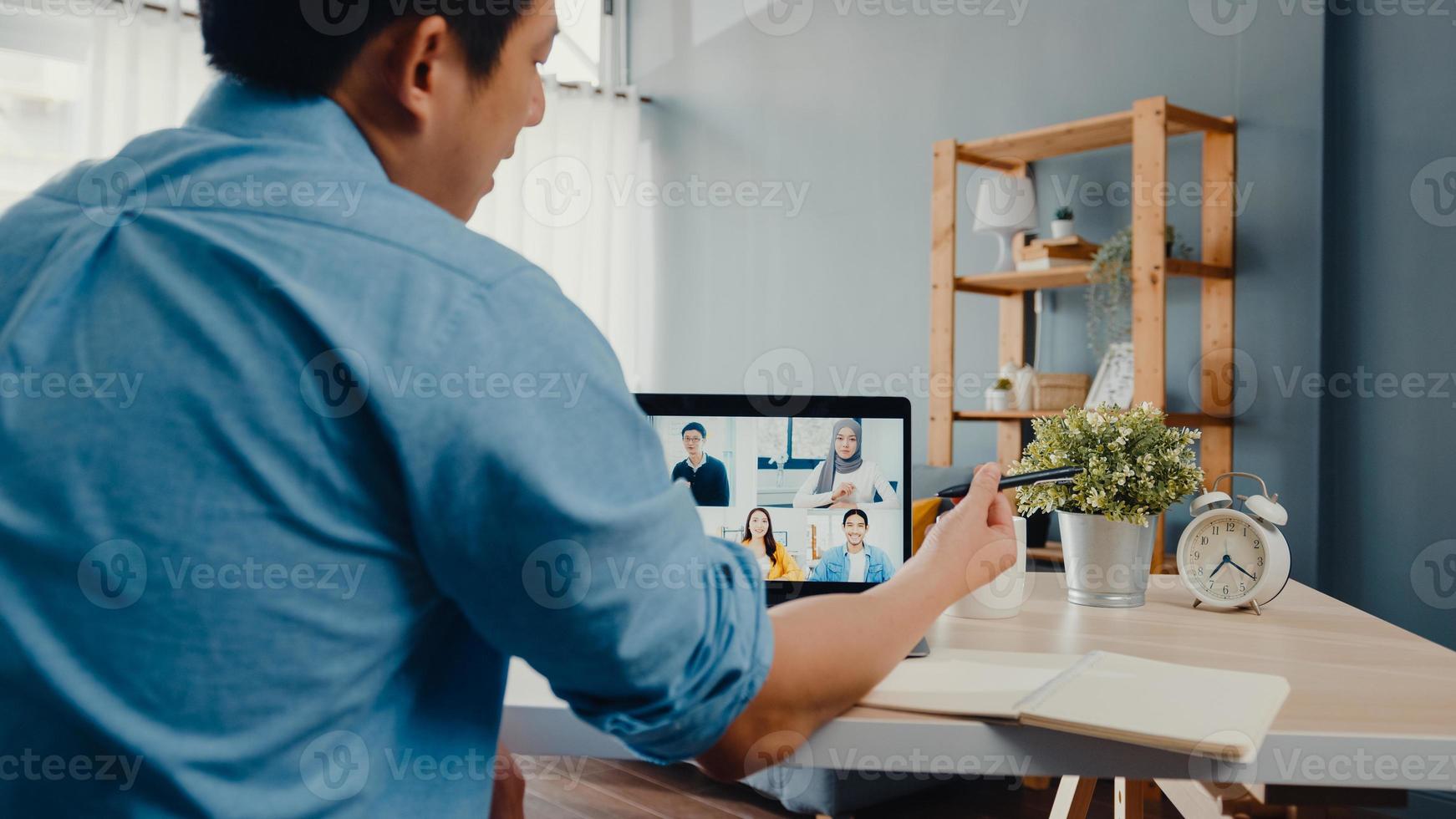 Young Asia businessman using laptop talk to colleagues about plan in video call meeting while work from home at living room. Self-isolation, social distancing, quarantine for corona virus prevention. photo