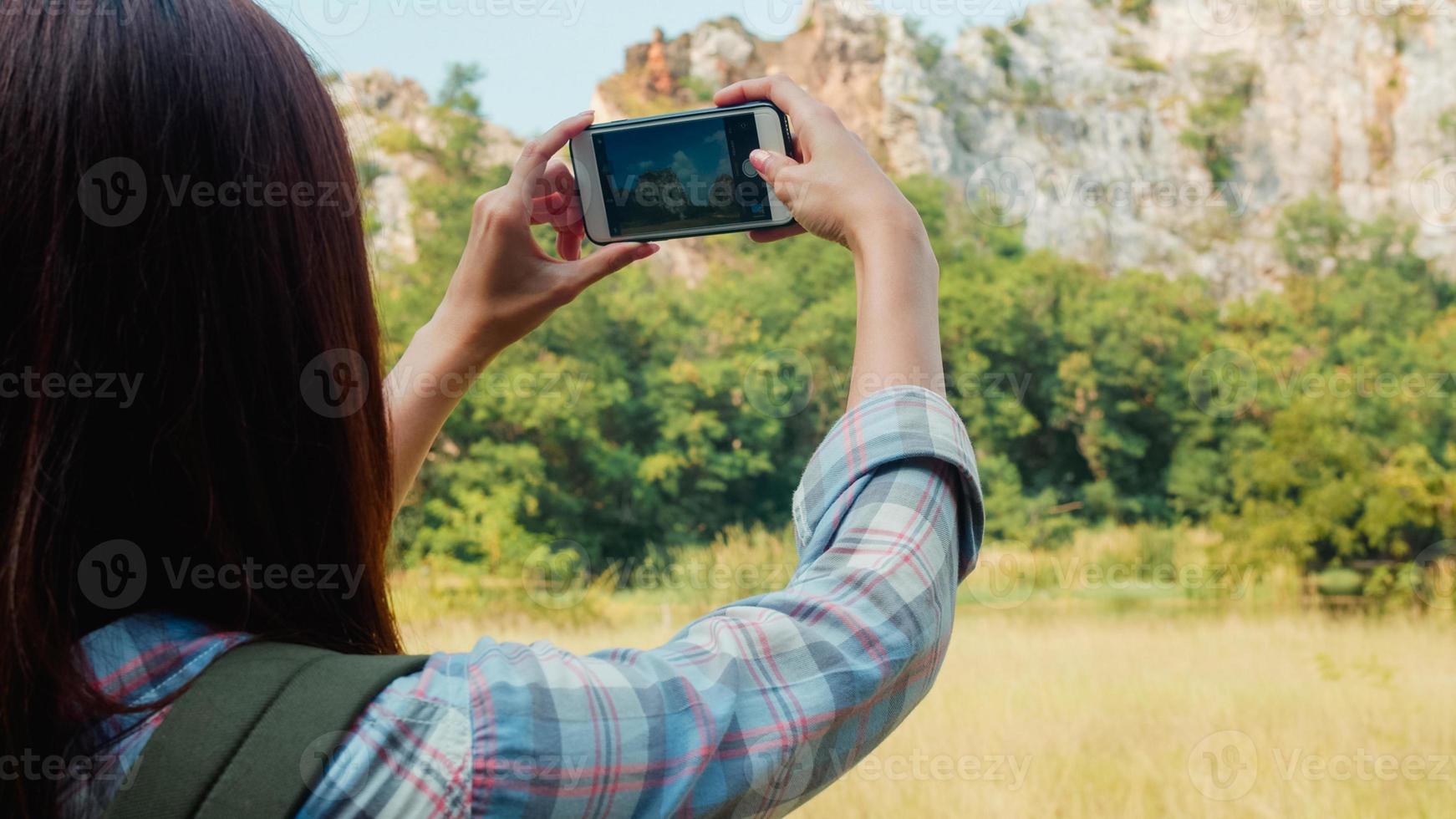 Cheerful young traveler Asian lady with backpack using smartphone for take a picture at mountain lake. Korean girl enjoy holidays adventure feeling happy freedom. Lifestyle travel and relax concept. photo