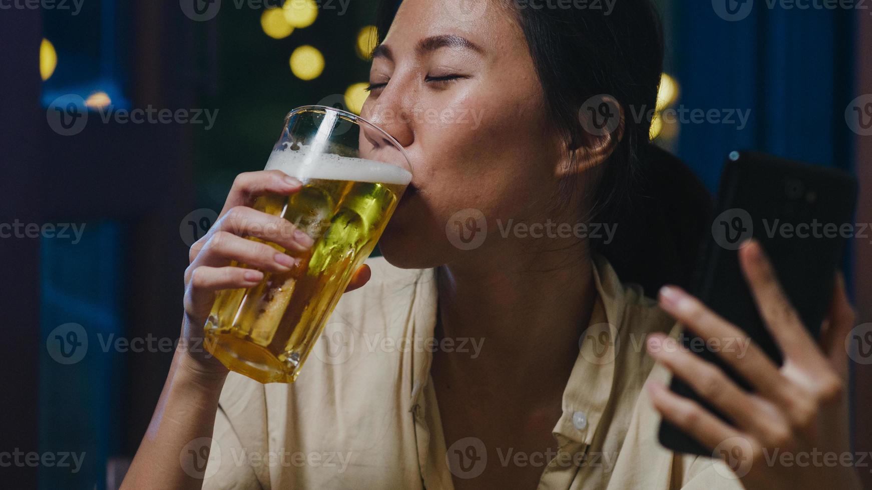 Young Asia lady drinking beer having fun happy moment night party New Year event online celebration via video call by phone at home at night. Social distancing, quarantine for coronavirus prevention. photo