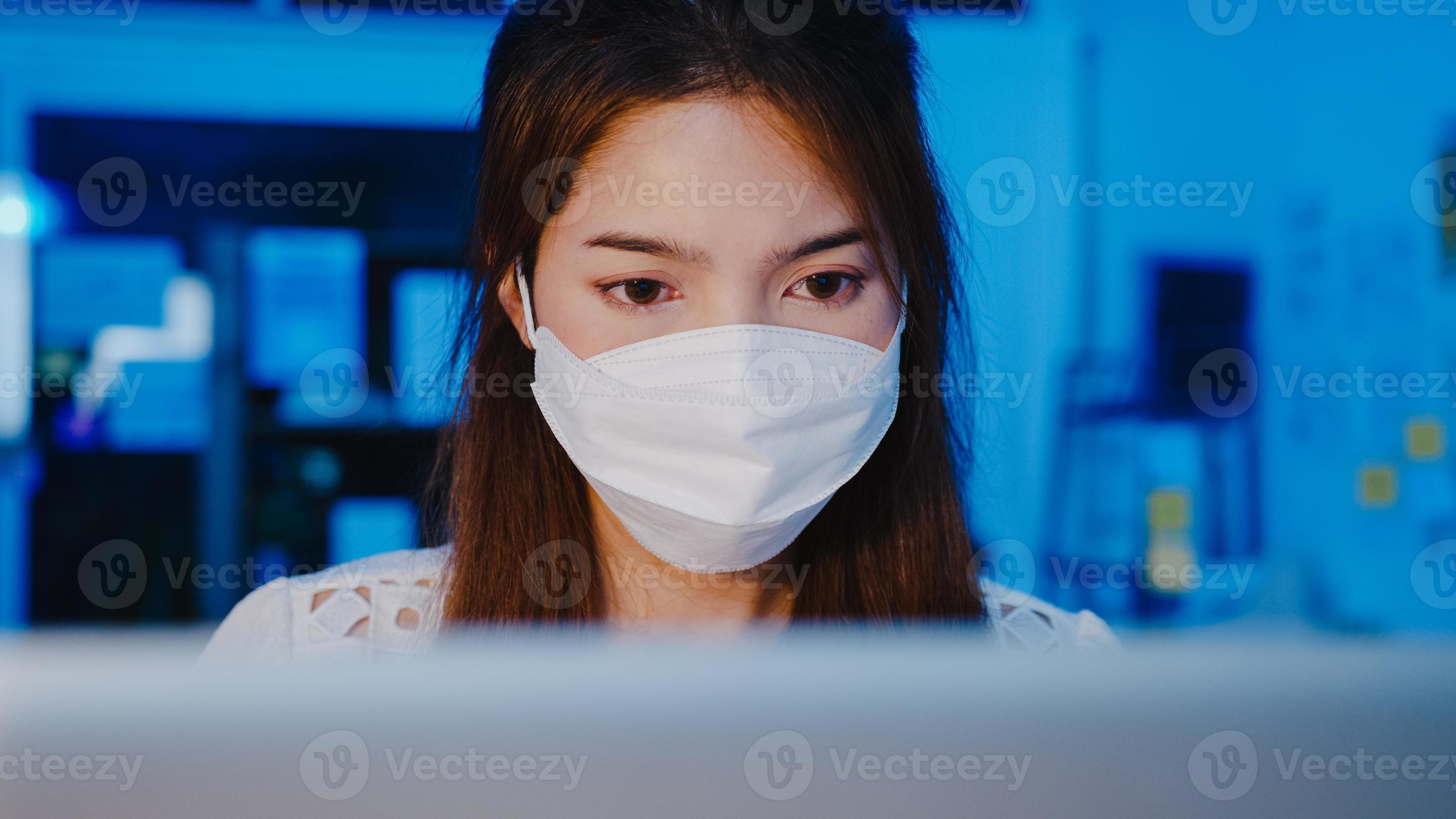 Happy Asia businesswoman wearing medical face mask for social distancing in new normal situation for virus prevention while using laptop back at work in office night. Life and work after coronavirus. photo